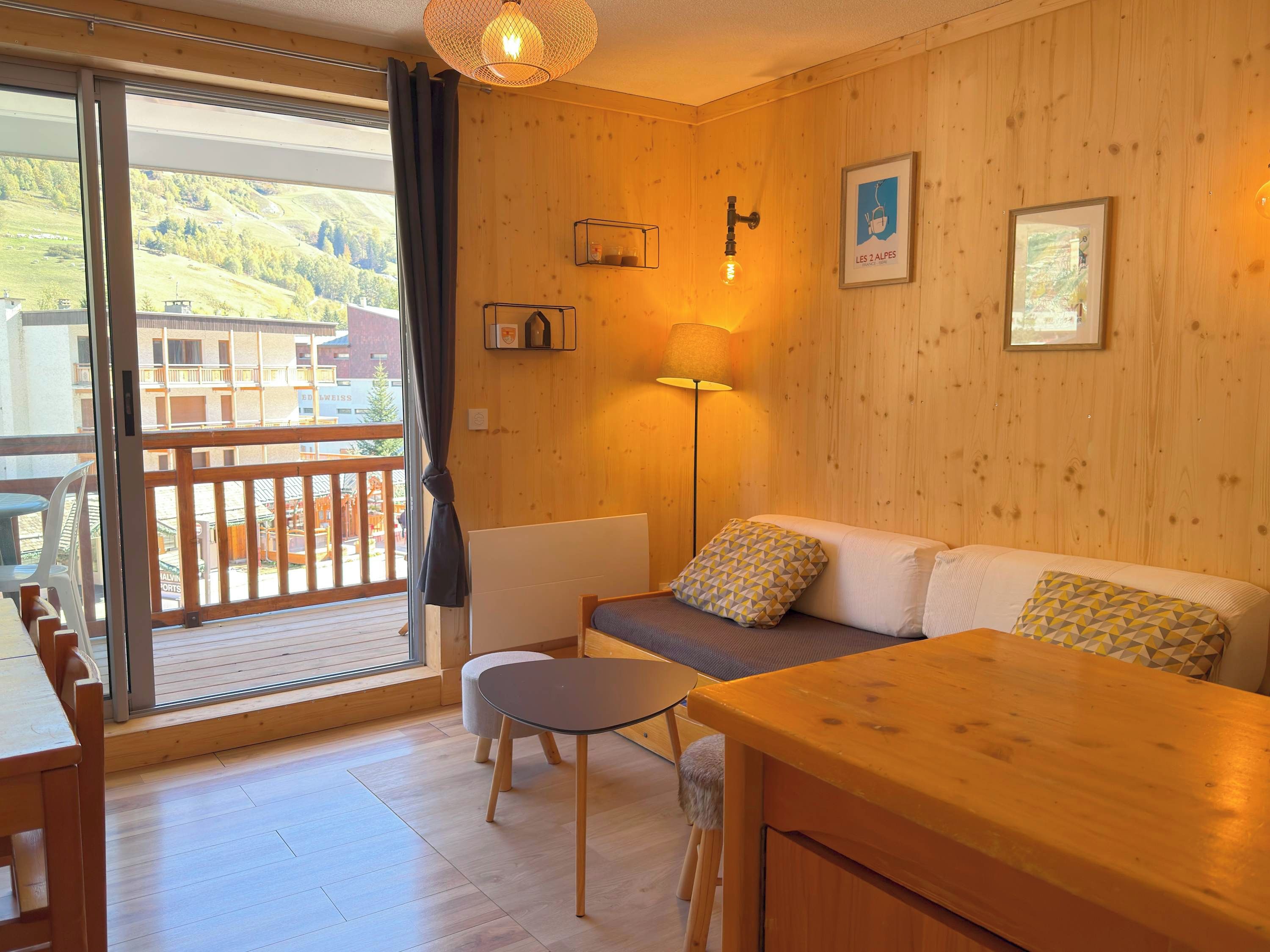 A cozy room with a wooden floor, a sofa, a small table, and a chair. There is a balcony with a view of the outside.