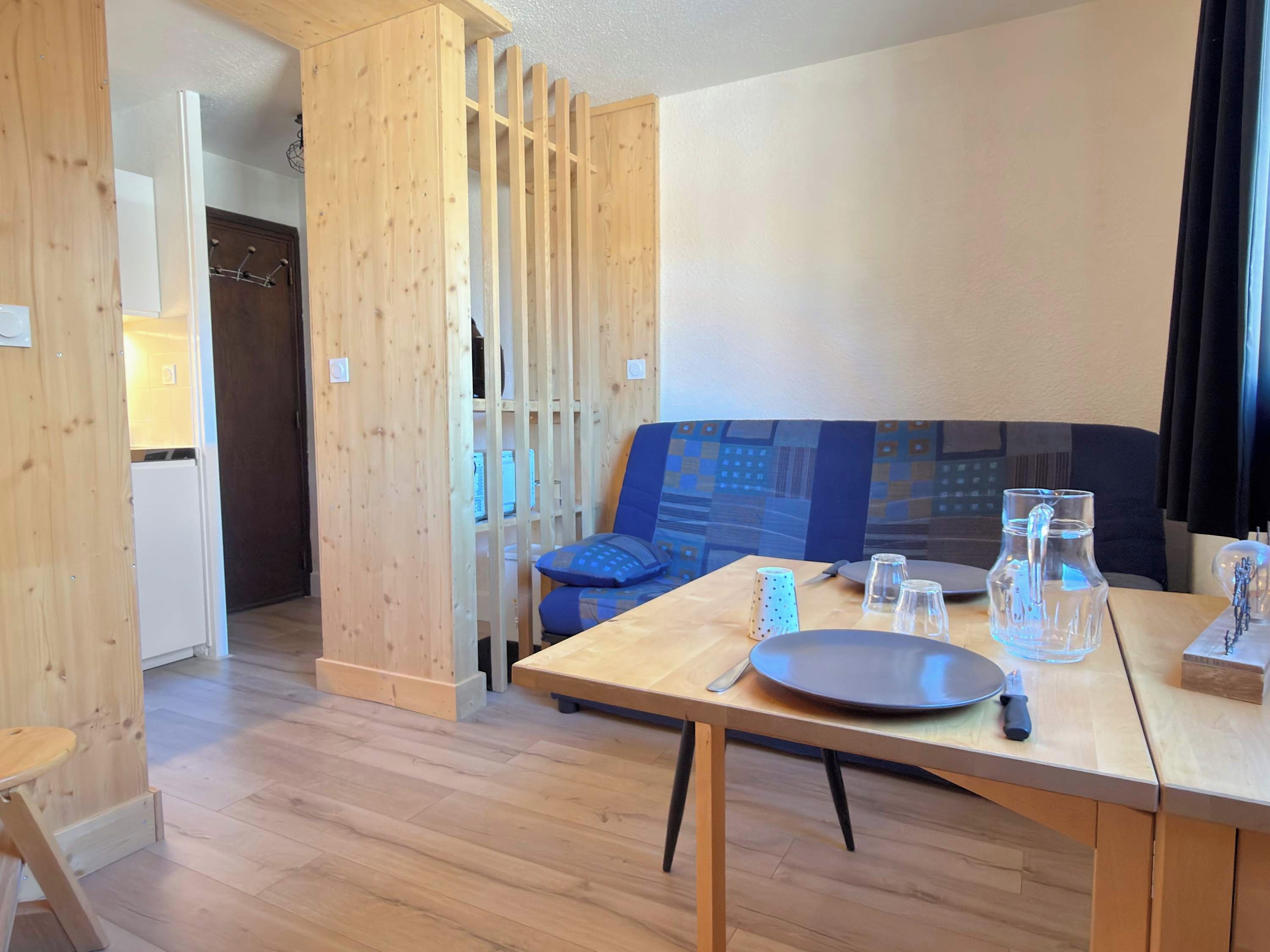 A modern room with a wooden floor, a dining table with chairs, a sofa, and a kitchen area visible through a wooden partition.