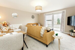 A living room with a sofa, a dining table set for two, a small side table, a television, and a large window with curtains. - The Forest Mews - 3 Bed Cottage Dalbeattie (Dumfries and Galloway)