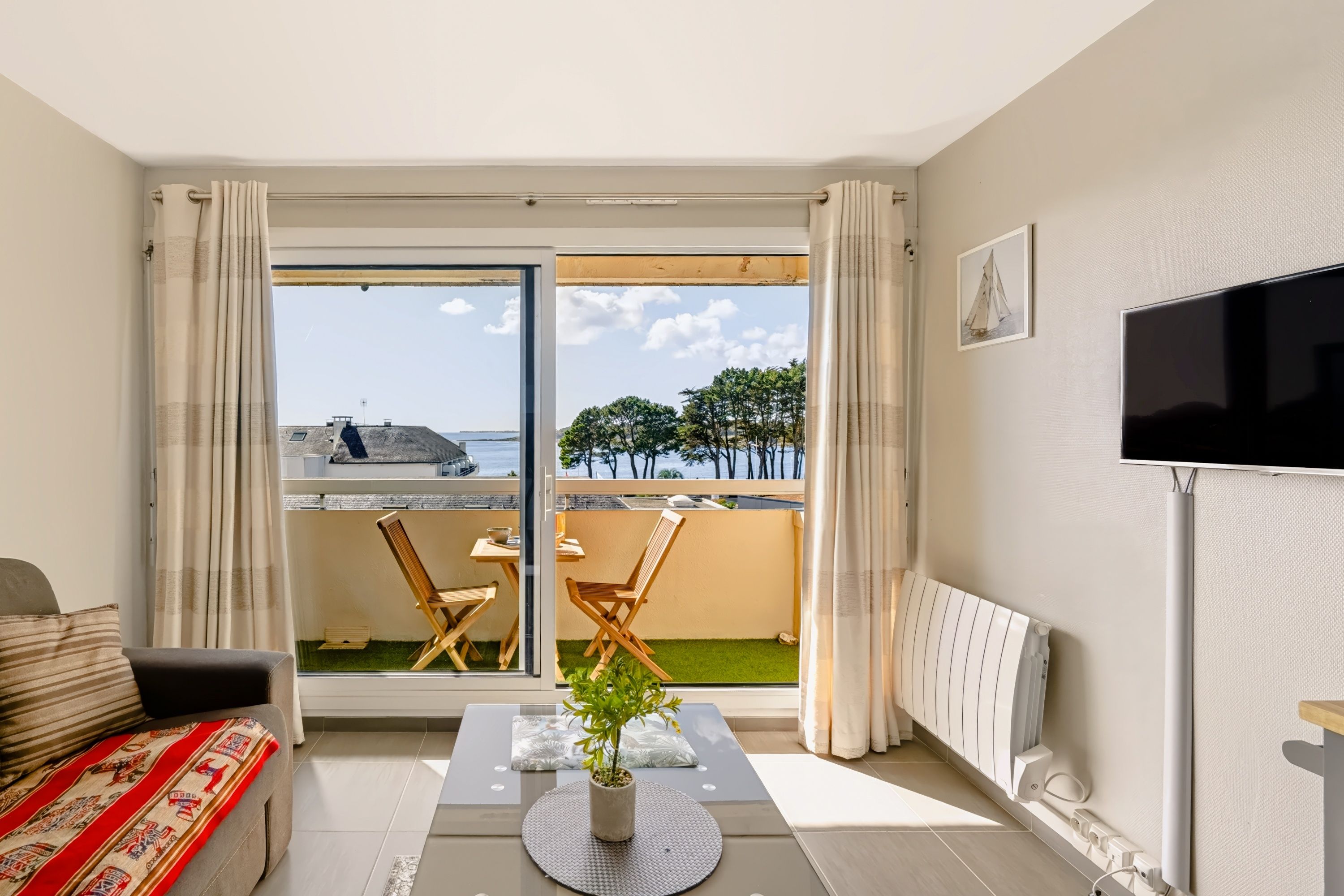 Ein modernes Wohnzimmer mit einer Couch, einem Esstisch und Stühlen, einem Fernseher und Meerblick durch große Glasfenster.