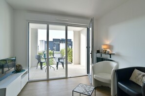 Ein modernes Wohnzimmer mit einer Couch, einem kleinen Tisch und einem Sessel. Eine Schiebetür aus Glas führt in einen Außenbereich mit Tisch und Stühlen.