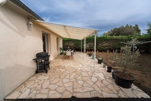 Un patio con suelo de piedra, una parrilla, una mesa con sillas y plantas en macetas.