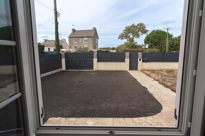 A view through a window of a paved outdoor area with a black surface, surrounded by a low wall and a gate. - La Maison entre 2 Caps - 2km de Fréhel (Plurien)