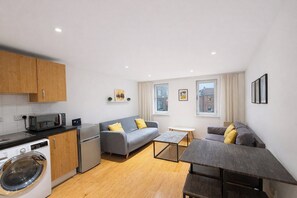 A compact living space with a kitchenette, a grey sofa with yellow cushions, a small table, and a washing machine. - Top Rated Prime Marylebone 1BR Well-Equipped for 3 (NEW) (London)