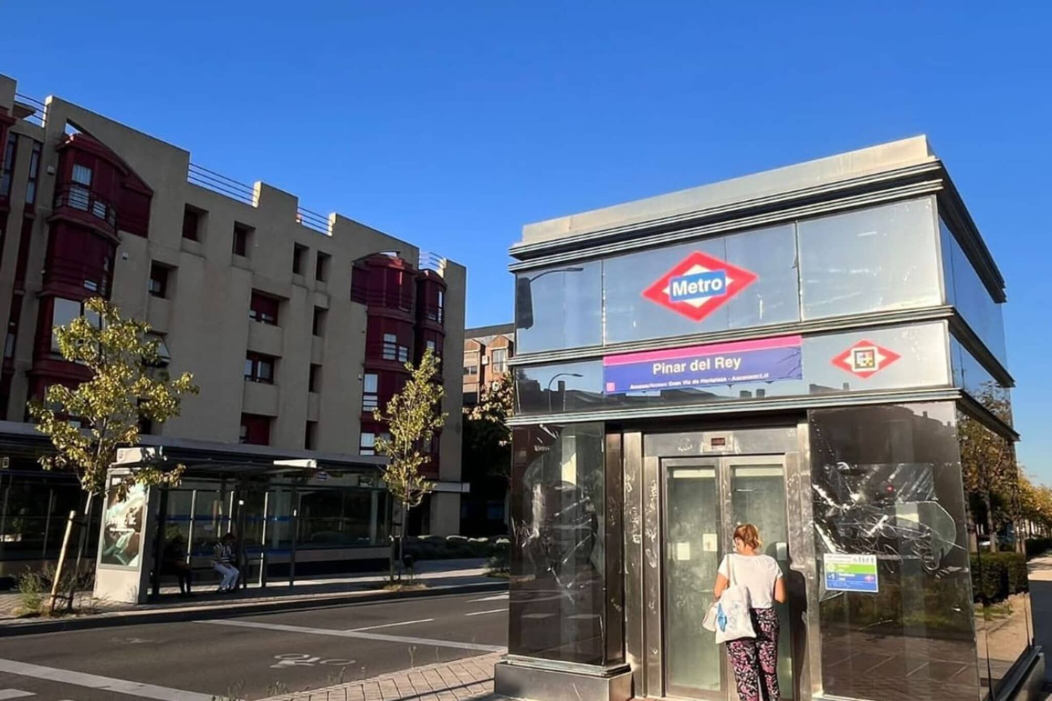 Una estación de Metro con cielo despejado, un refugio de autobús y un edificio con fachada roja y blanca.