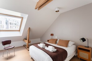 Een moderne slaapkamer met een schuine kap, een bed met bruin en wit beddengoed, een houten stoel, een nachtkastje met een lamp, en uitzicht op een gebouw door een raam.