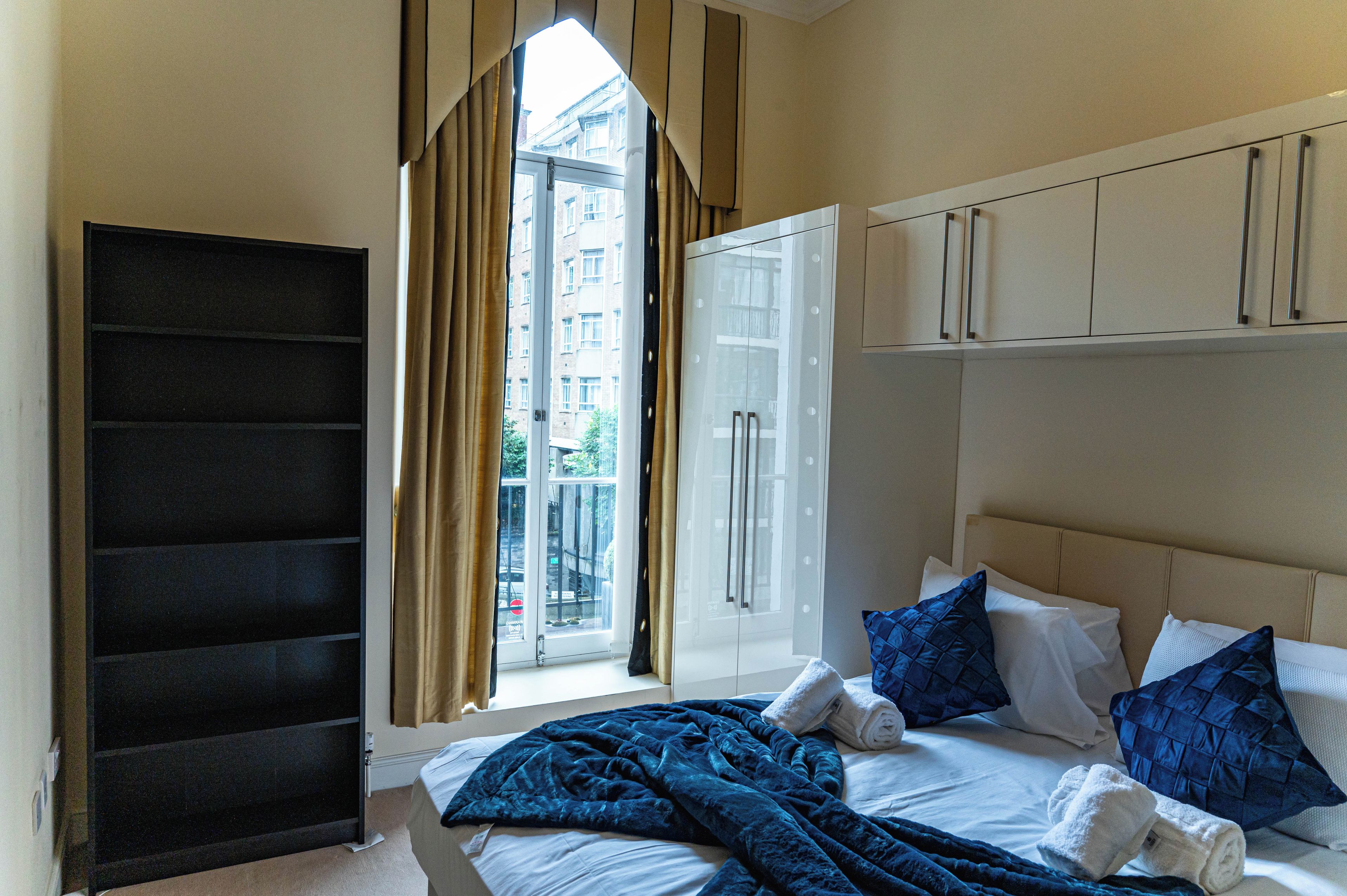 A hotel room with a bed, a window with curtains, a wardrobe, and a view of buildings outside.