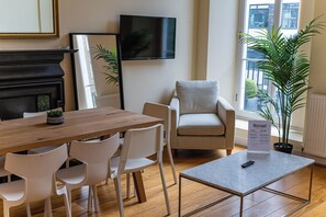 A modern living room with a wooden dining table, white chairs, a grey armchair, a flat-screen TV mounted on the wall, and a potted plant. - The Russel Square Apartmens (London)