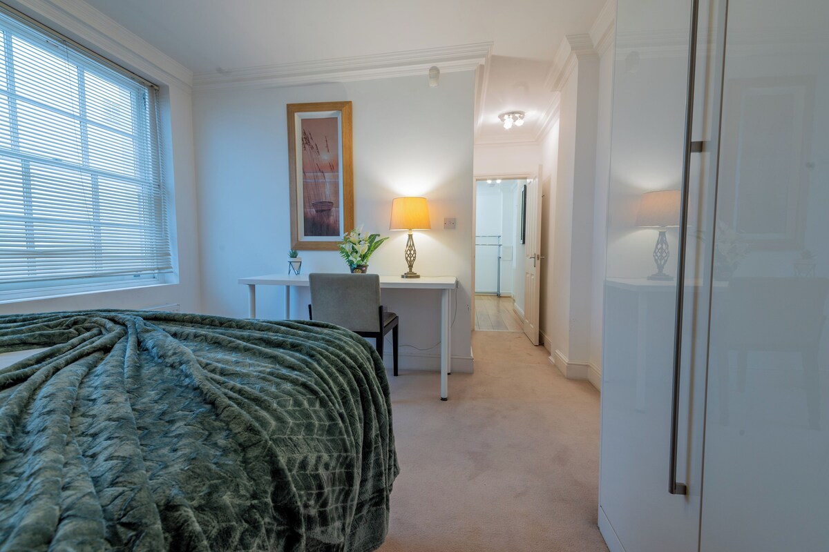 A bedroom with a bed, a desk, and a chair. There is a window with blinds, a lamp, and a framed picture on the wall.
