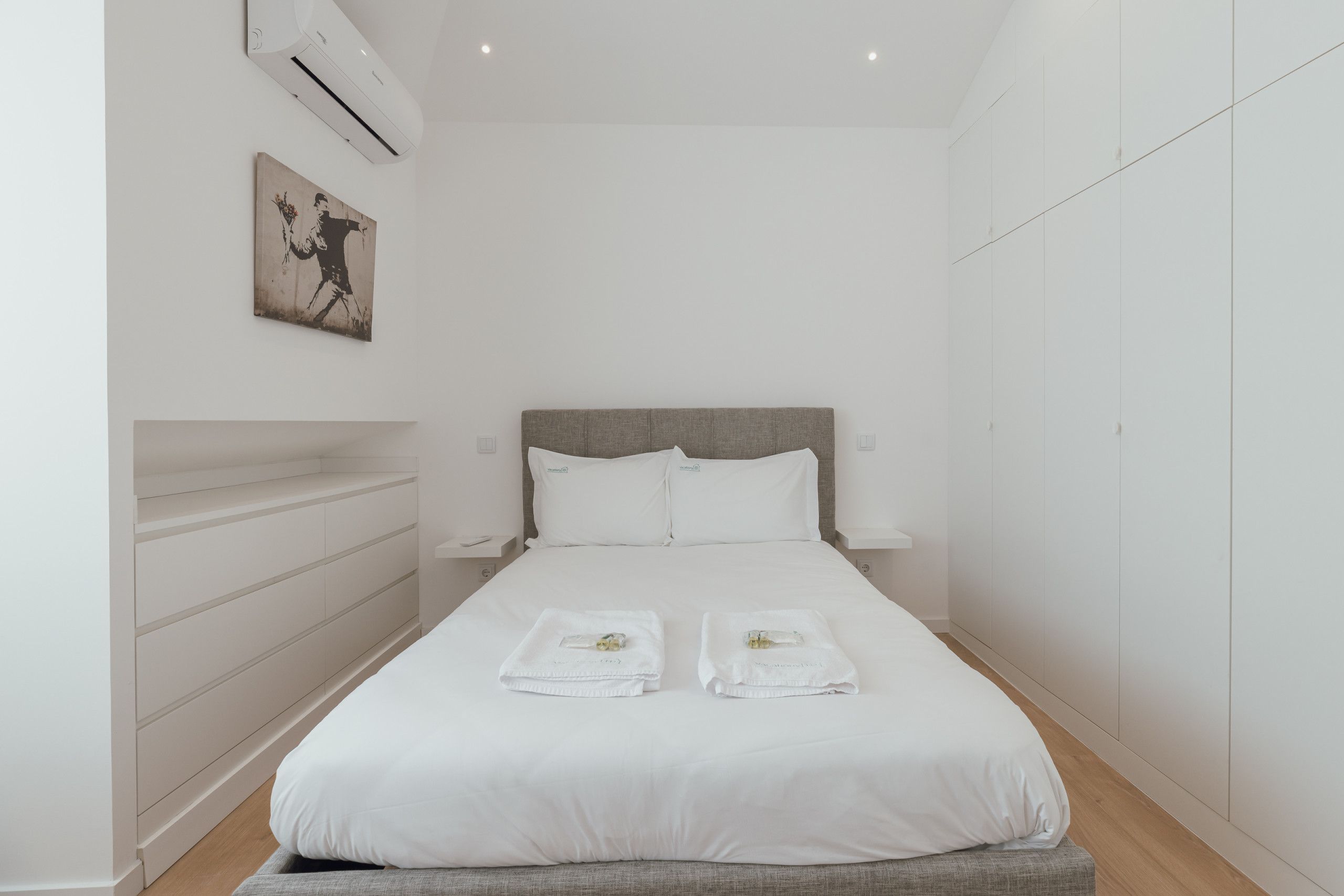 Een minimalistisch hotelkamer met een groot bed, witte lakens, een grijs hoofdeinde, en één kunstwerk aan de muur.