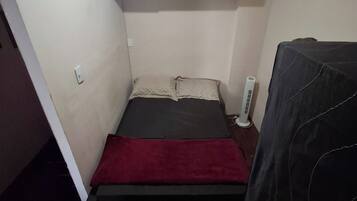 Une petite pièce à la lumière tamisée, avec un lit, un ventilateur de plafond et une étagère fixée au mur, où sont rangés des livres.