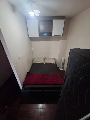 A small, dimly lit room with a bed, a ceiling fan, and a wall-mounted shelf with books.