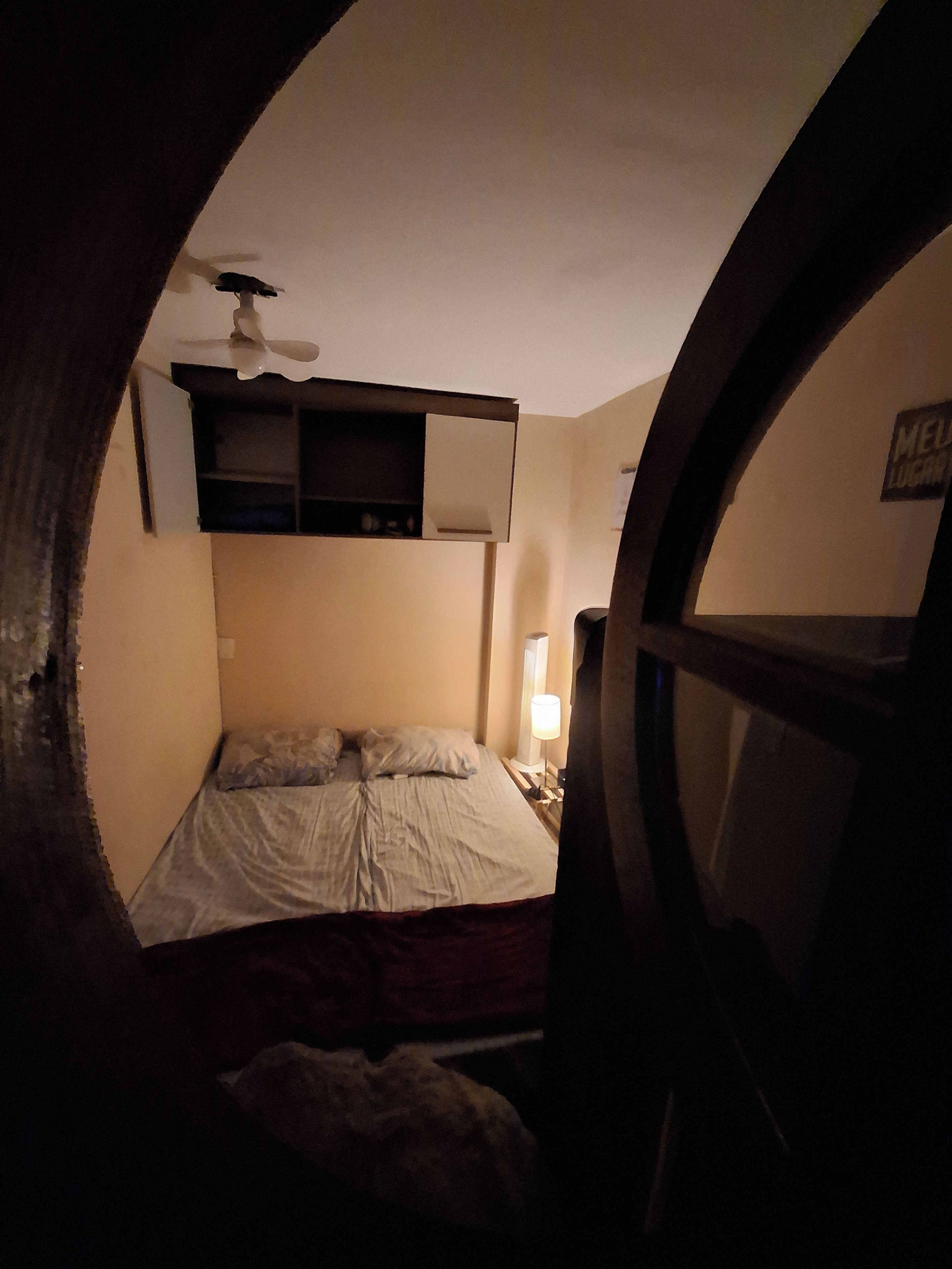 A dimly lit room with a bed, a ceiling fan, and a wall-mounted shelf.