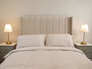 A bed with a striped bedspread, two bedside tables with lamps, and a padded headboard. - Long Meadow Apartment (Ballymena)