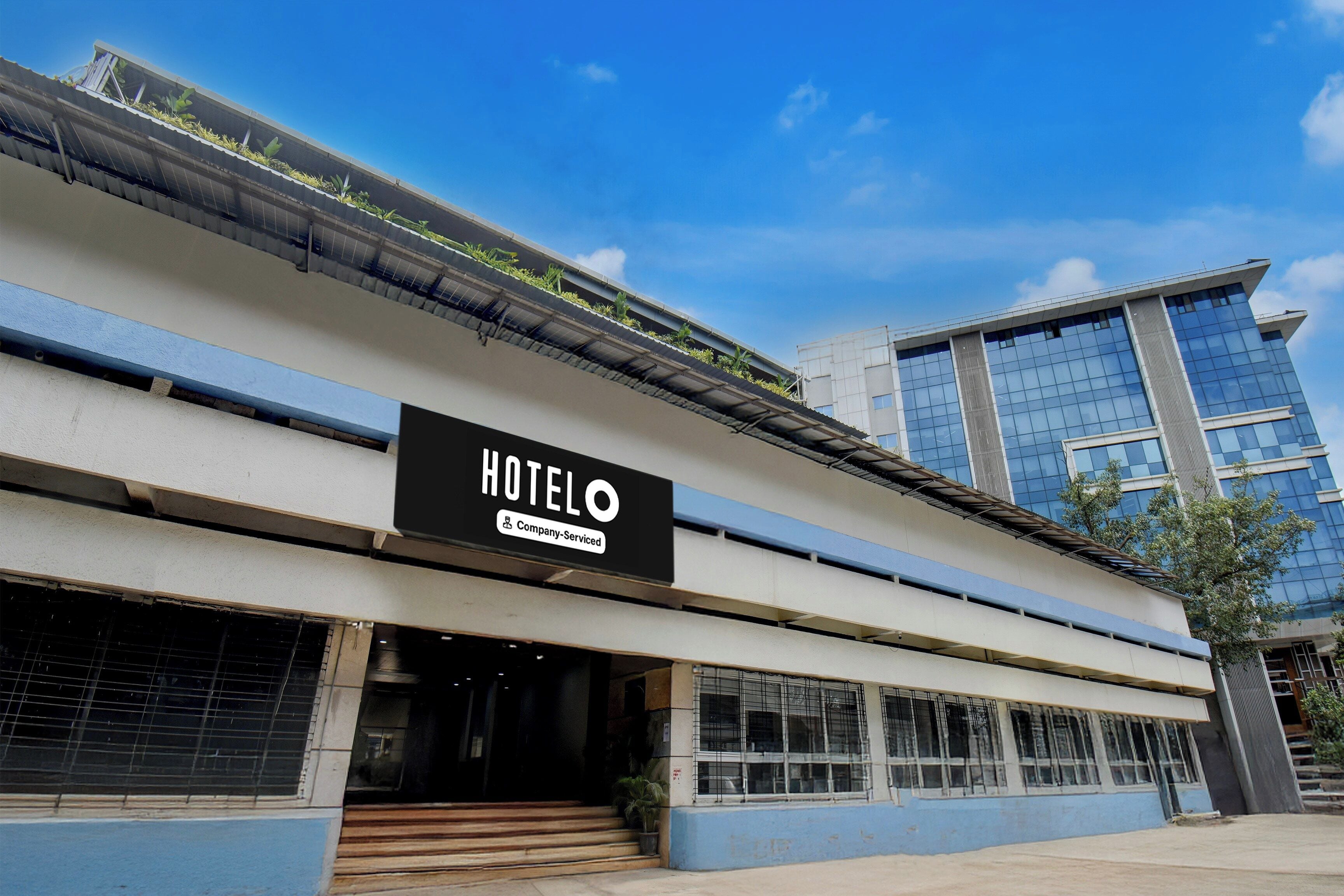 Un hôtel avec une enseigne « HOTELO » bien visible, un logo indiquant qu’il est affilié à une chaîne, et un bâtiment moderne avec de grandes fenêtres en arrière-plan.