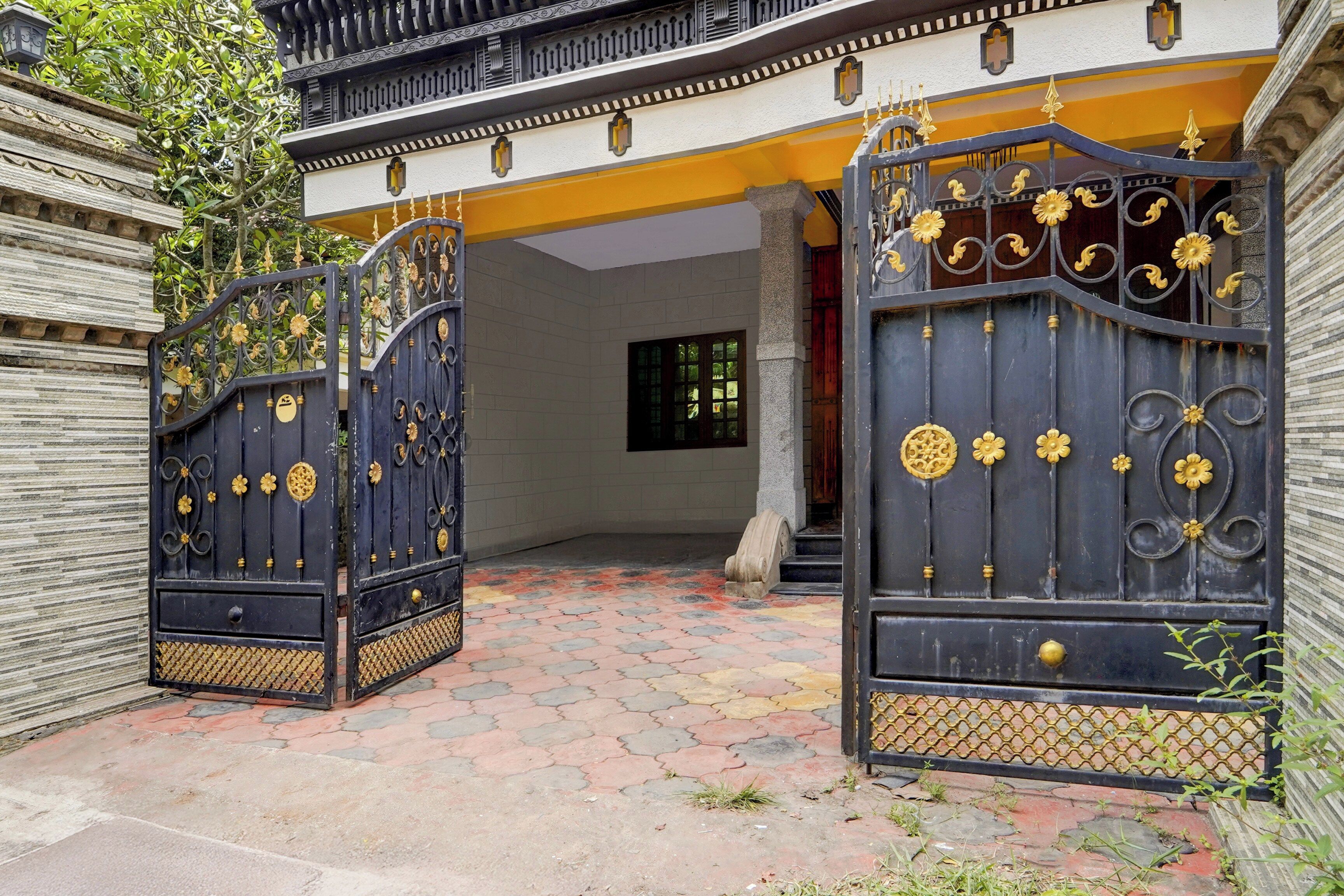 A gated entrance with decorative metalwork and a paved pathway leading to a building with a yellow facade and a window.