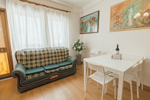 Ein Wohnzimmer mit einem Karomuster-Sofa, einem Esstisch mit Stühlen, einer Topfpflanze und zwei Bildern an der Wand.