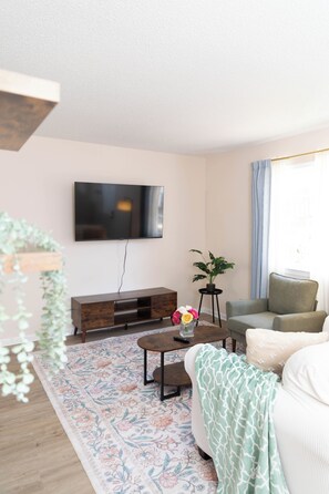 Ein Wohnzimmer mit einem an der Wand befestigten Flachbildfernseher, einer Holzkommode, einem Couchtisch mit Blumenvase, einer Couch und einem Fenster mit Vorhängen.