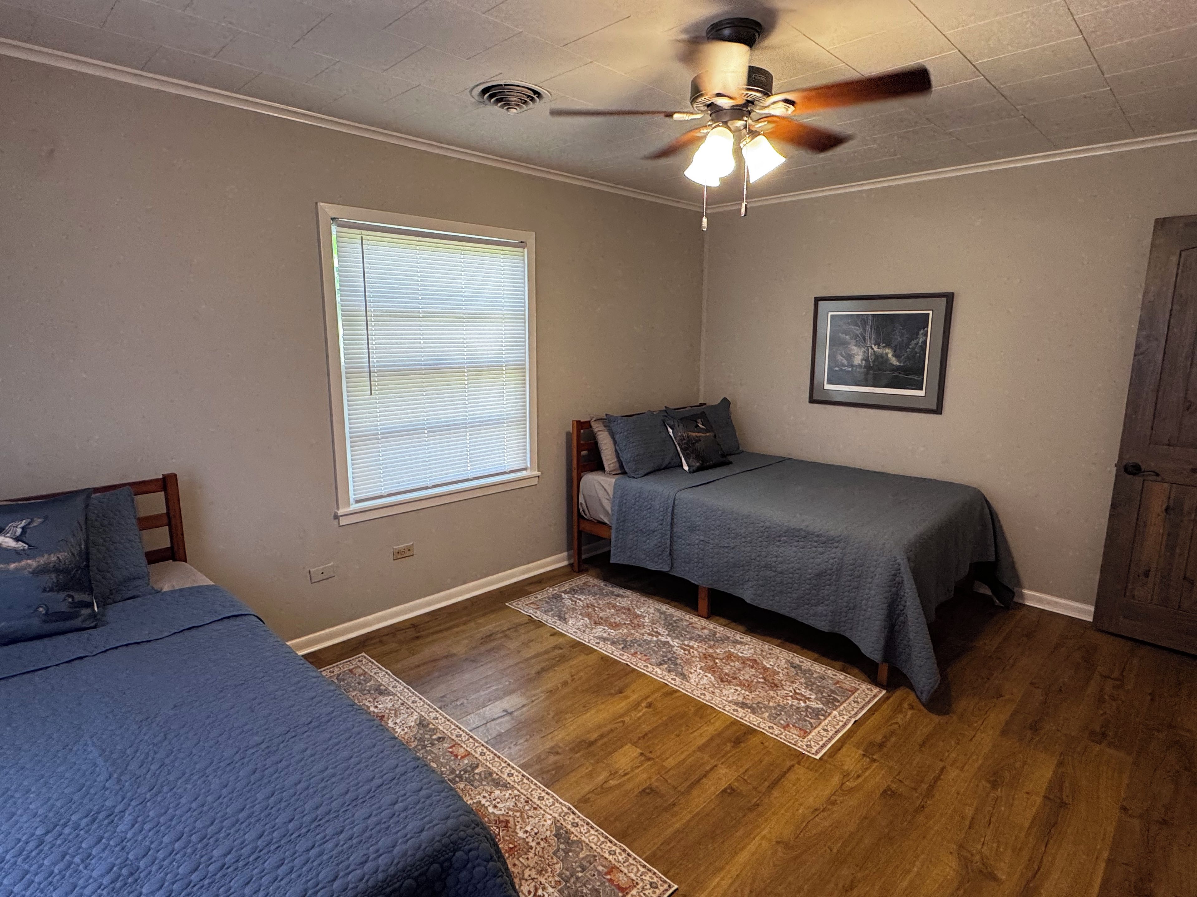 A bedroom with two beds, a ceiling fan, a window with blinds, a rug, and a framed picture on the wall.