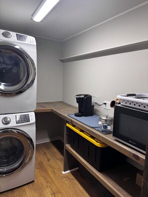 A laundry room with stacked washing machines, a coffee maker, a microwave, and a small table with various items.