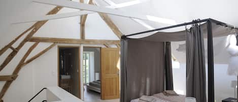 Une chambre avec un plafond haut, des poutres apparentes, un lit à baldaquin, une bibliothèque et une vue sur l’extérieur.