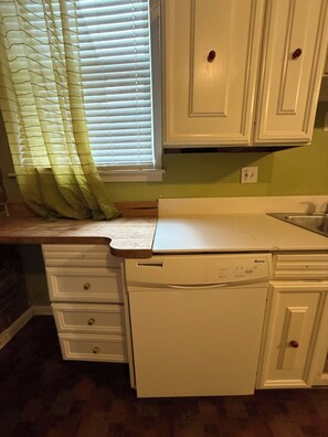 A kitchen with white cabinets, a dishwasher, and a window with blinds. - Row House Suite 927 (Topeka)