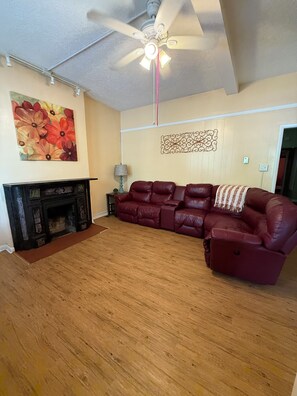A living room with a fireplace, a red leather sofa, a ceiling fan, and a floral painting on the wall.