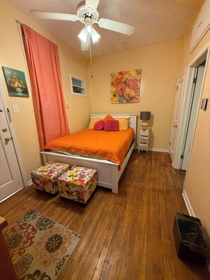 A bedroom with a bed, a ceiling fan, a nightstand, a lamp, a window with curtains, and a door. - Row House Suite 927 (Topeka)