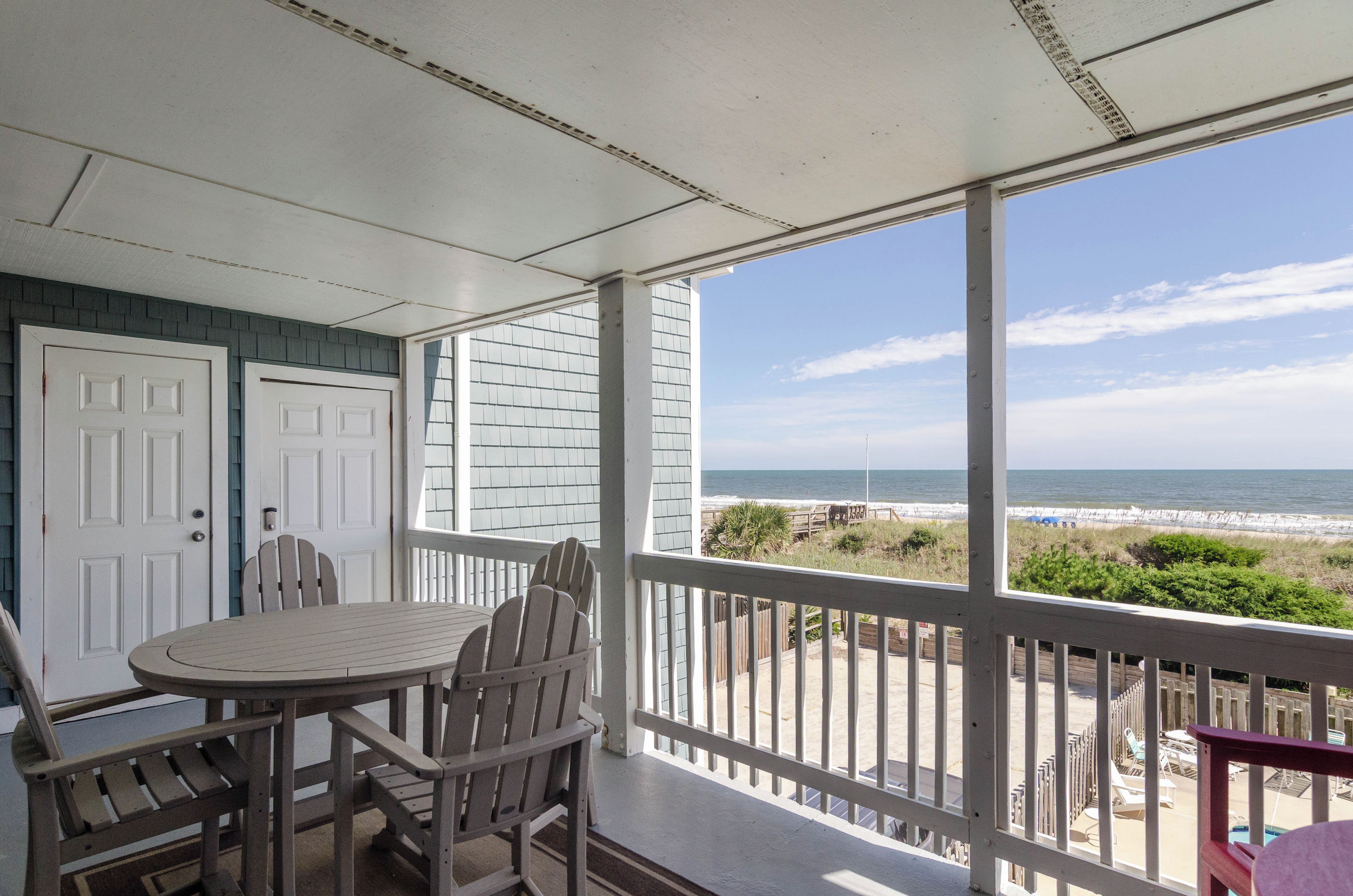 A covered balcony with a table and chairs, overlooking a beach with a wooden fence and lounge chairs.