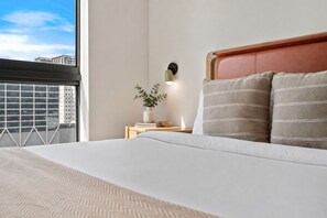 A hotel room with a large bed, a nightstand with a plant, and a view of buildings through a window. - The Crosby 2BR Balcony by Palermo Home (Miami)