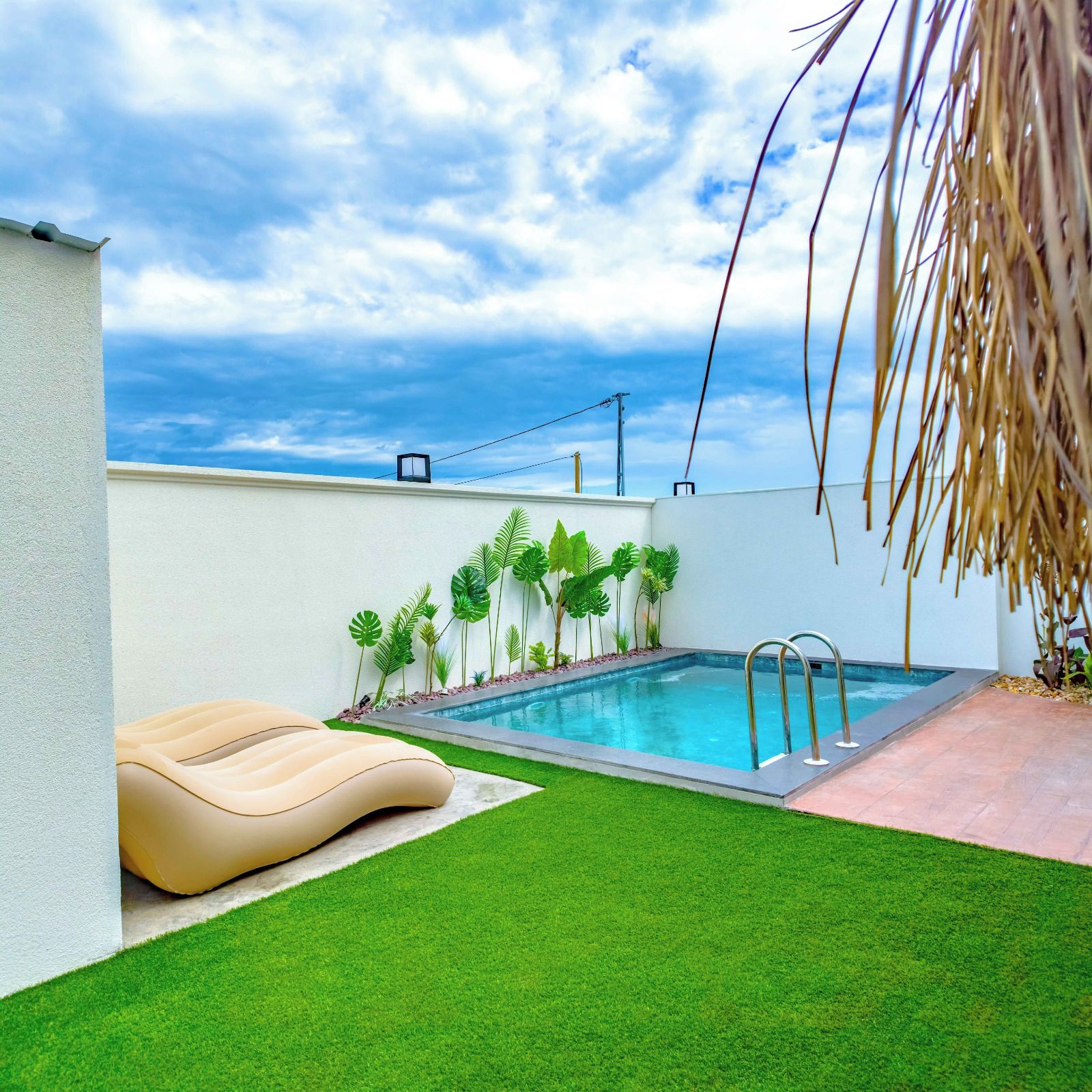 A swimming pool with a metal ladder, surrounded by artificial grass and tropical plants.