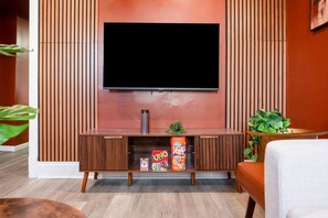 A modern living room with a flat-screen TV mounted on a wooden panel wall, a wooden entertainment unit with a small plant and snacks, and a comfortable seating area with a white sofa. - Comfy 2 Bedroom - Games - 1 Garage Spot - Sleeps 6 (Chicago)