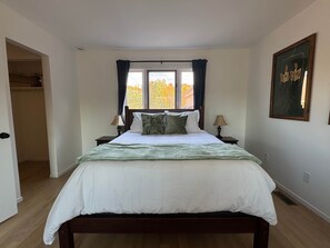 Une chambre à coucher avec un lit, deux tables de chevet, une fenêtre avec des rideaux, une porte et un tableau encadré au mur.