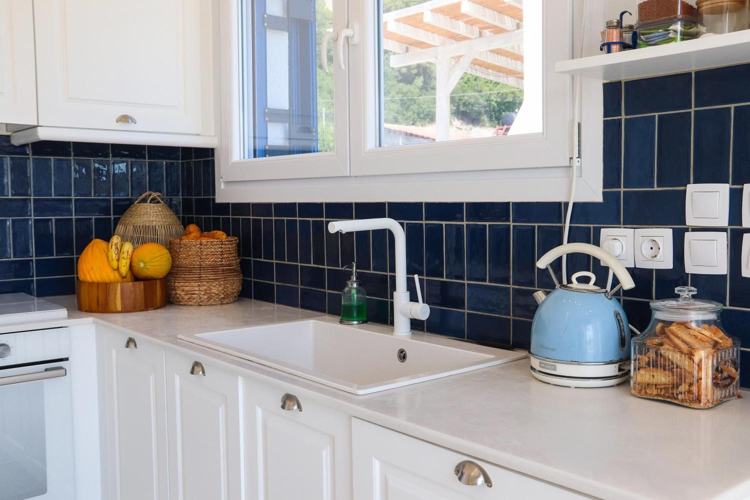 A kitchen with a white sink, a blue kettle, and a window above the sink.