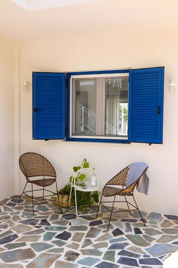 Een kamer met een betegelde vloer, twee rieten stoelen, een kleine tafel met een plant en een raam met blauwe luiken.