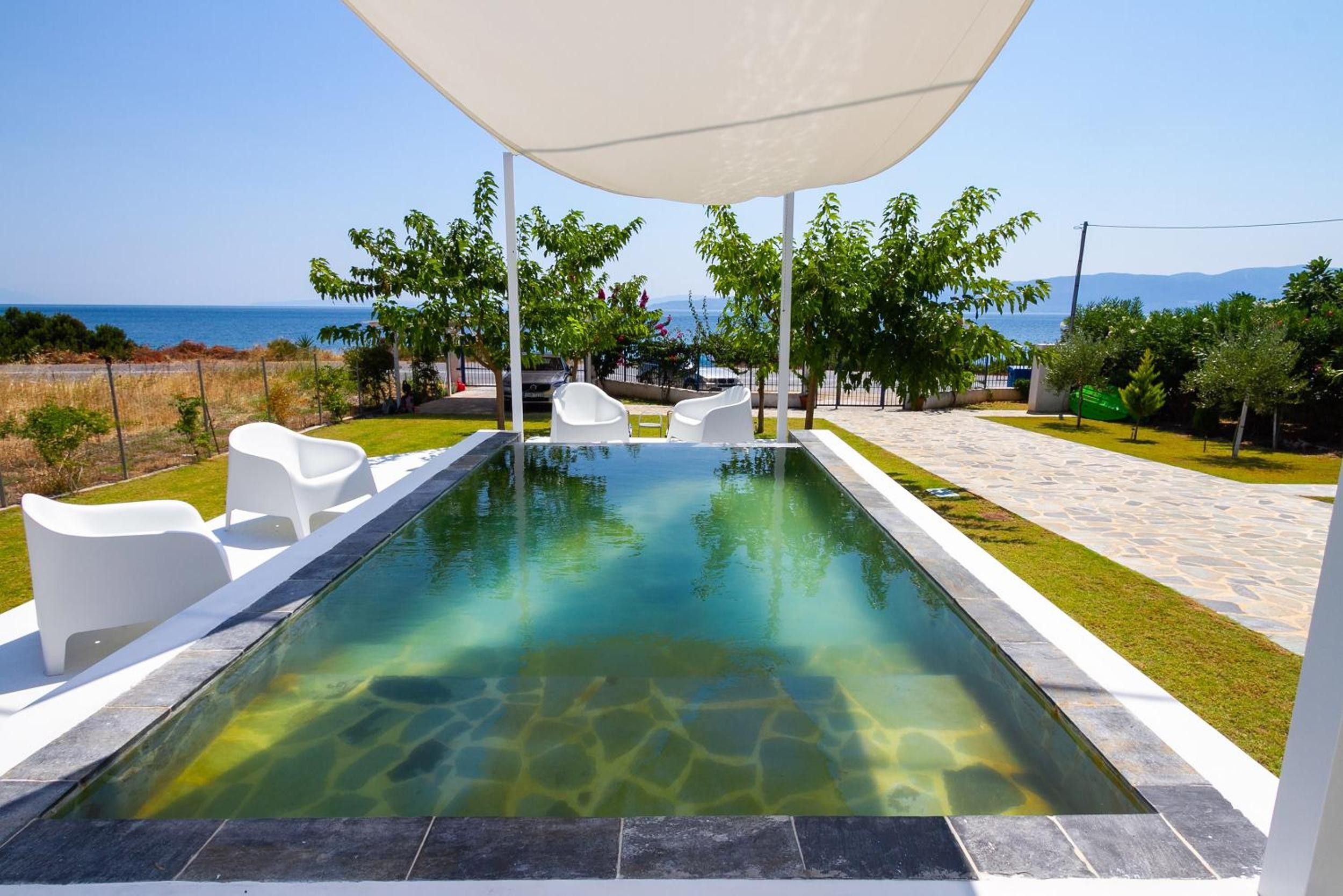 A swimming pool with clear water, surrounded by white lounge chairs and a canopy, with a view of the sea and mountains in the background.