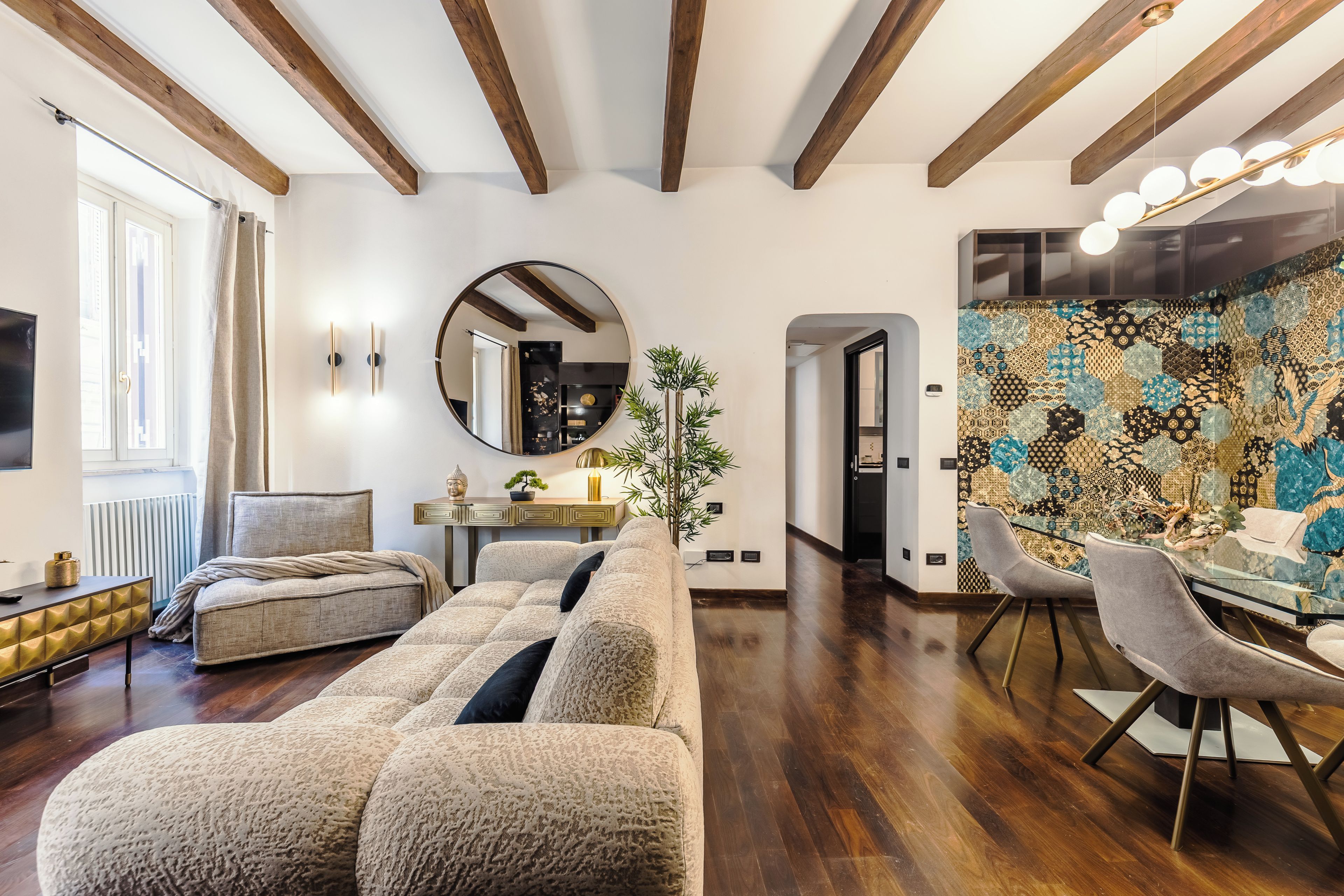 A modern living room with a sofa, a dining area with chairs, a round mirror, and a patterned wall.