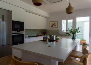 A modern kitchen with white cabinetry, a central island, stainless steel appliances, and a dining area with wicker chairs. - Casa Perdiz, ideal choice for style and comfort (Conil de la Frontera)