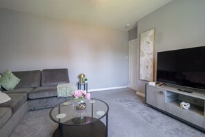 A living room with a grey sofa, a glass coffee table with flowers, a flat-screen TV on a stand, and a wall with a floral artwork. - Ground Floor 2 Bed Apt in Salsburgh (Salsburgh)