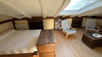 Un dormitorio con cama de madera, una mesita de noche de madera, una silla y una pequeña mesa. Hay un tragaluz sobre la cama.