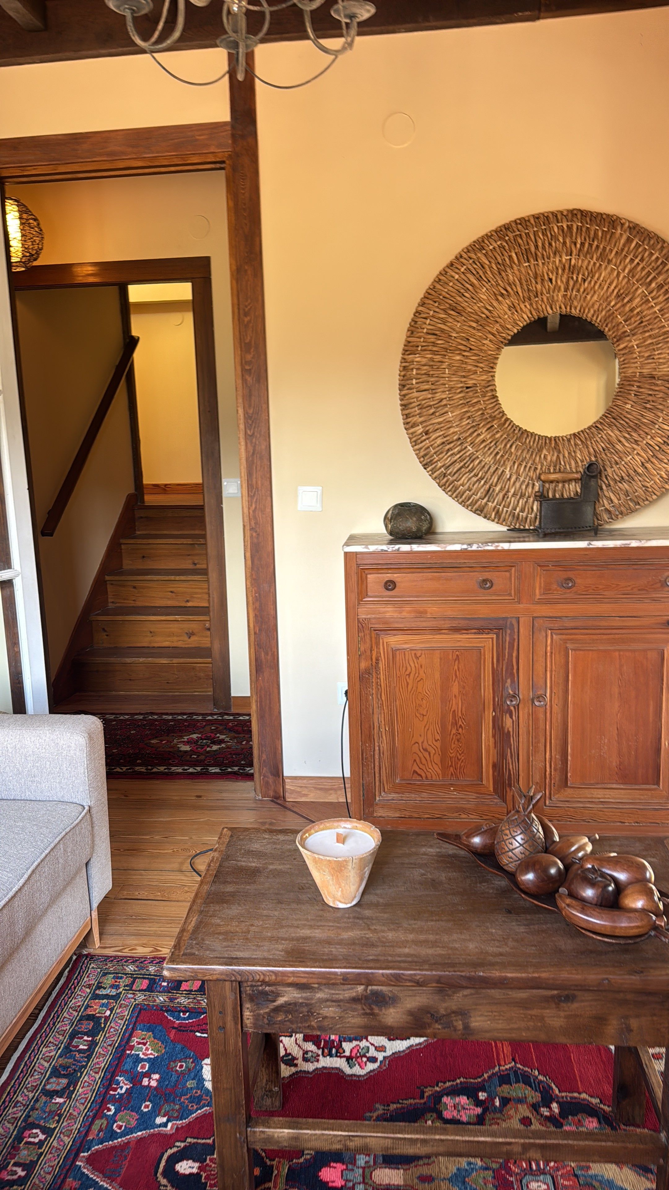 Un escalier en bois menant à un deuxième étage, un miroir rond en osier accroché au mur, un meuble en bois orné d’un vase, et un tapis à motifs sur le sol.