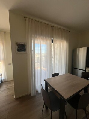 Un comedor con mesa y sillas, un refrigerador y un ventanal con cortinas.