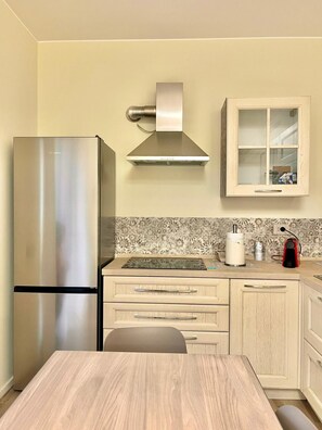 A kitchen with a stainless steel refrigerator, a range hood, a cabinet with glass doors, and a wooden table. - New Listing (Catania)