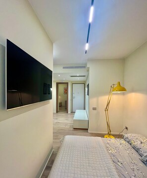 A modern hotel room with a bed, a desk, a flat-screen TV mounted on the wall, and a standing lamp. - New Listing (Catania)