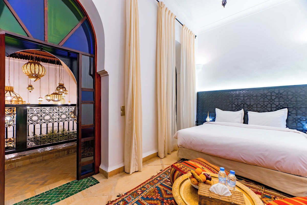 A hotel room with a bed, a decorative wall, a window with curtains, and a small table with a basket and bottles.