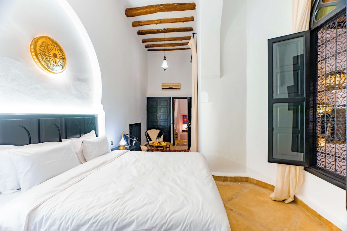 A bedroom with a large bed, a wooden ceiling, a small table with a chair, and a window with decorative iron bars.