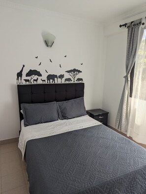 A bedroom with a bed, a nightstand, a wall-mounted lamp, and a window with curtains.