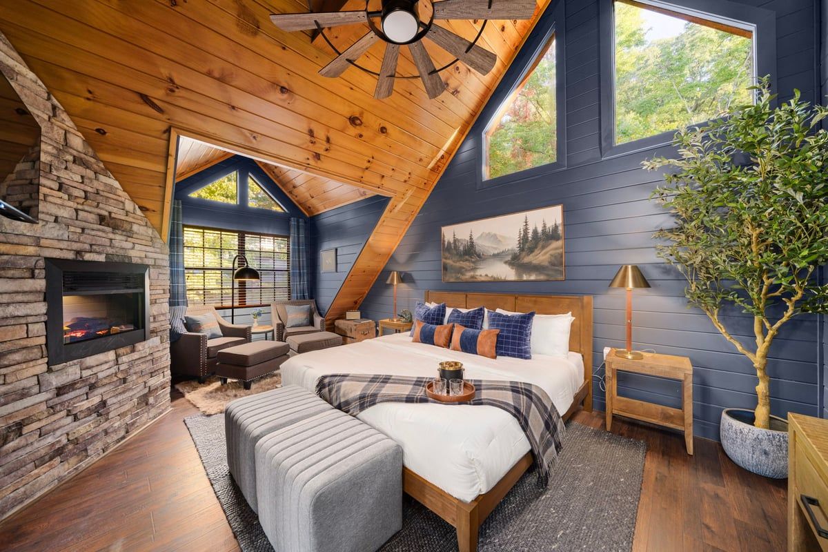 A bedroom with a stone fireplace, a large bed, a ceiling fan, and a view of the outdoors.