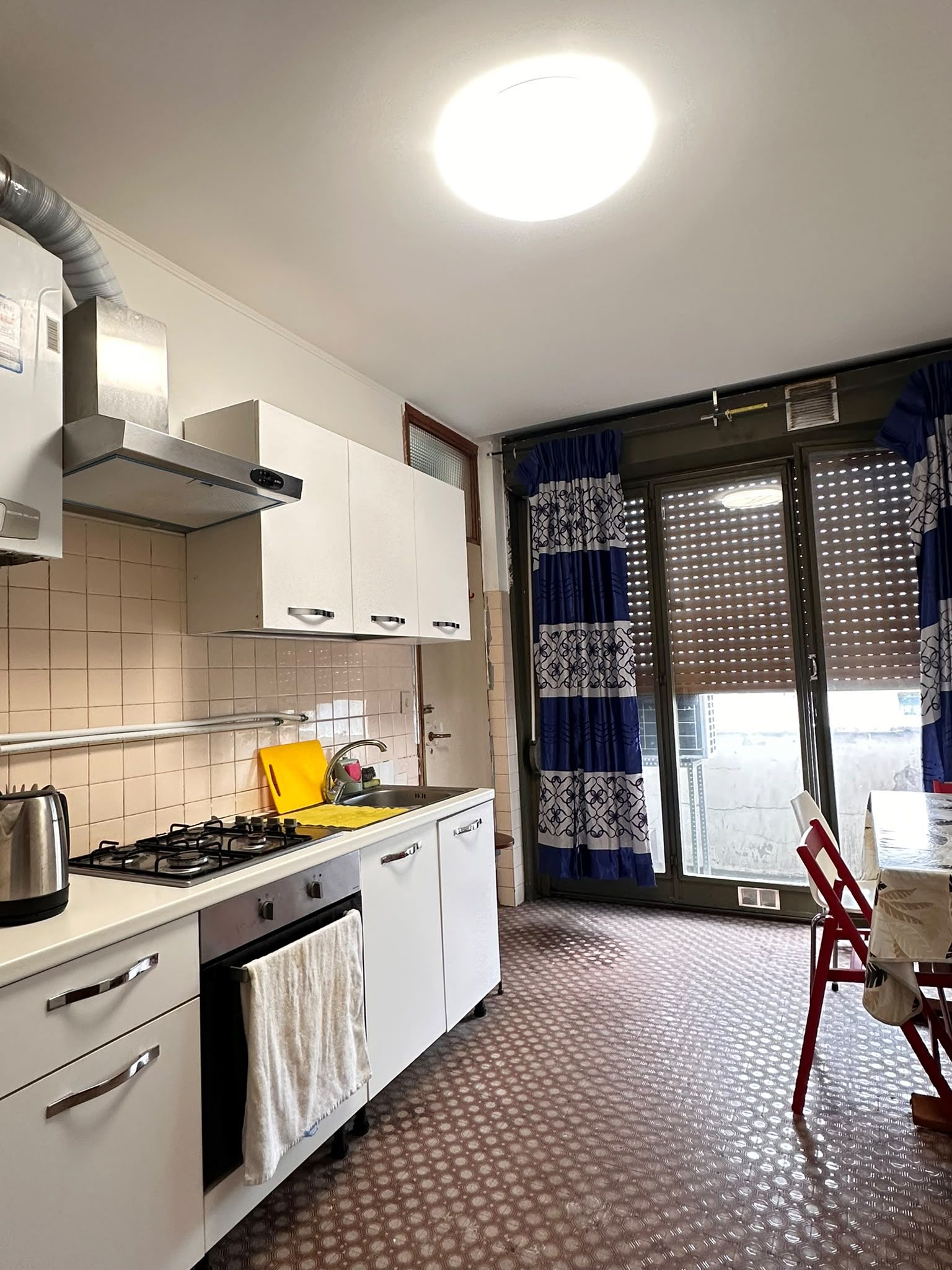 A kitchen with a gas stove, a sink, and cabinets. There is a window with blue curtains and a chair outside.