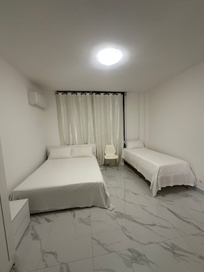 A room with two beds, a chair, a window with curtains, and an air conditioning unit. - New Listing (Venezia)